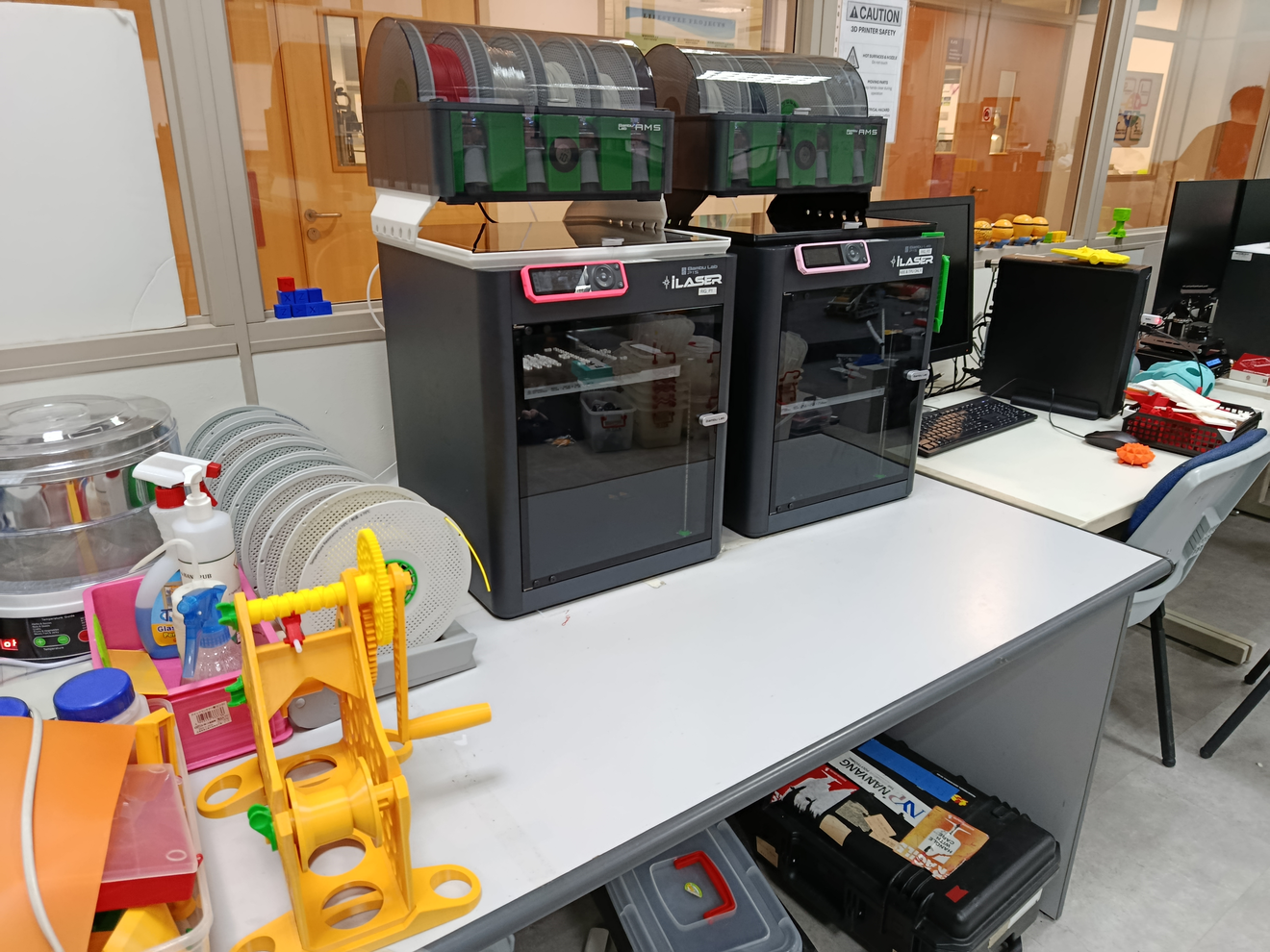 A workshop setting featuring two industrial-grade 3D printers lined up on a white table. The printers are black and prominent. 
