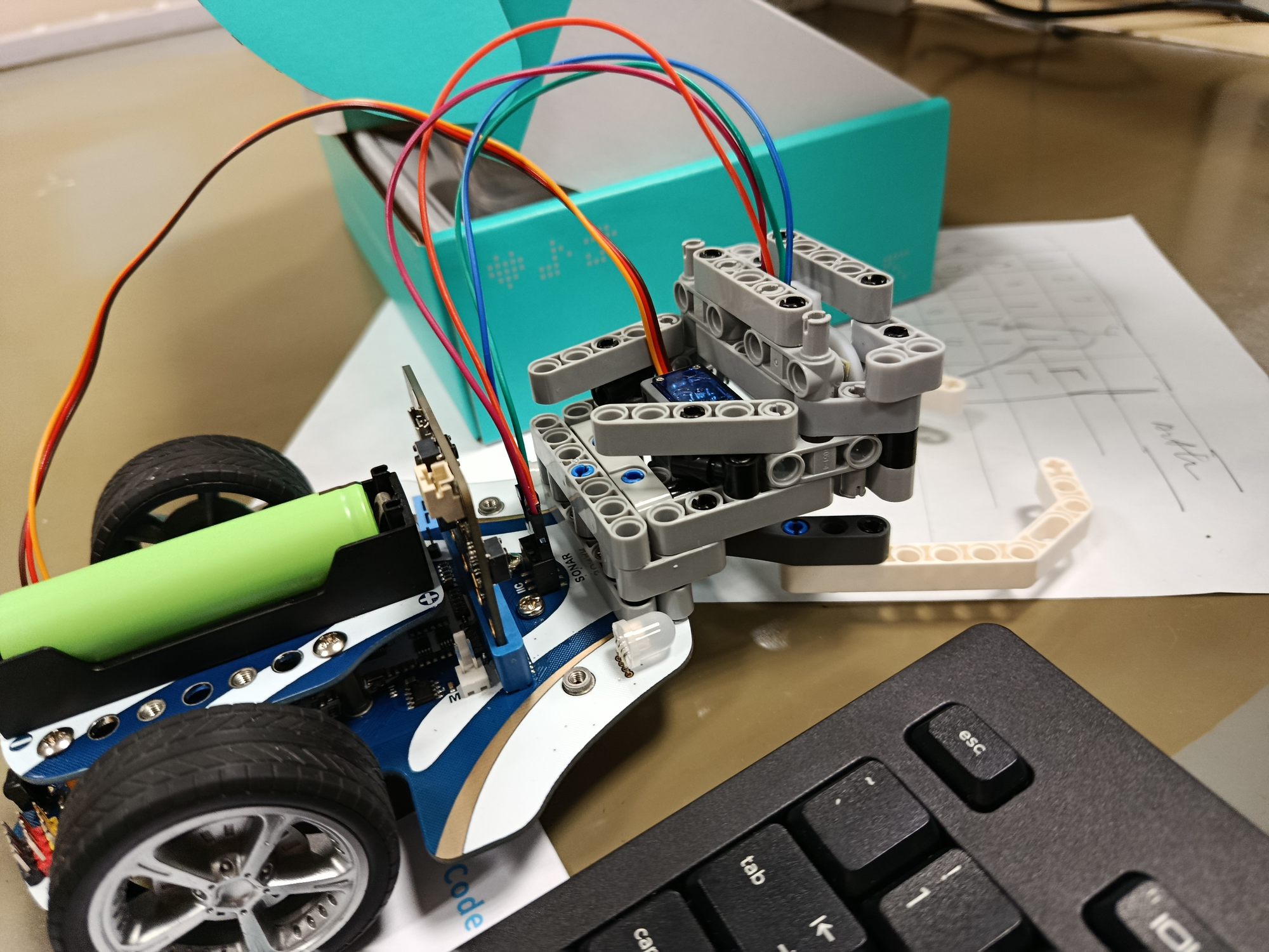 A close-up photo of a small, partially assembled robotic prototype. The robot is constructed from various electronic components, visible wiring (red, blue, orange), small motors, and a green-and-white chassis body.
