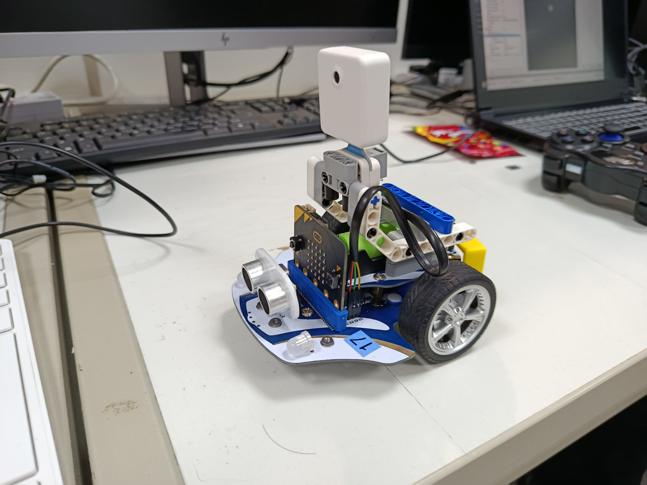 A detailed photo of a small, DIY robotic prototype resting on a white desk. The robot features a visible chassis, multiple electrical components, and wiring, including a central microcontroller board and two driven wheels.