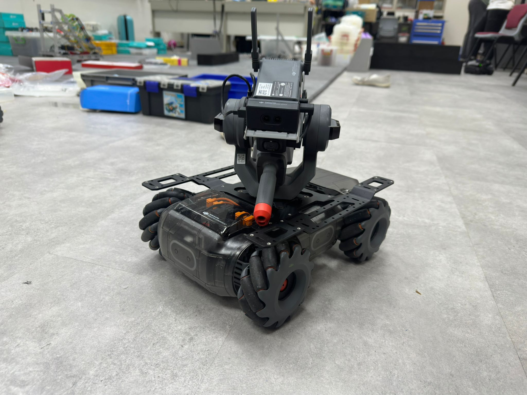 A photo of a tracked robotic prototype, sitting on a light-colored floor in a workshop environment. The robot has a large, black chassis and uses motorized treads for movement. Mounted on top of the chassis is an electronic assembly featuring a white vertical mount, various sensors, and wiring.
