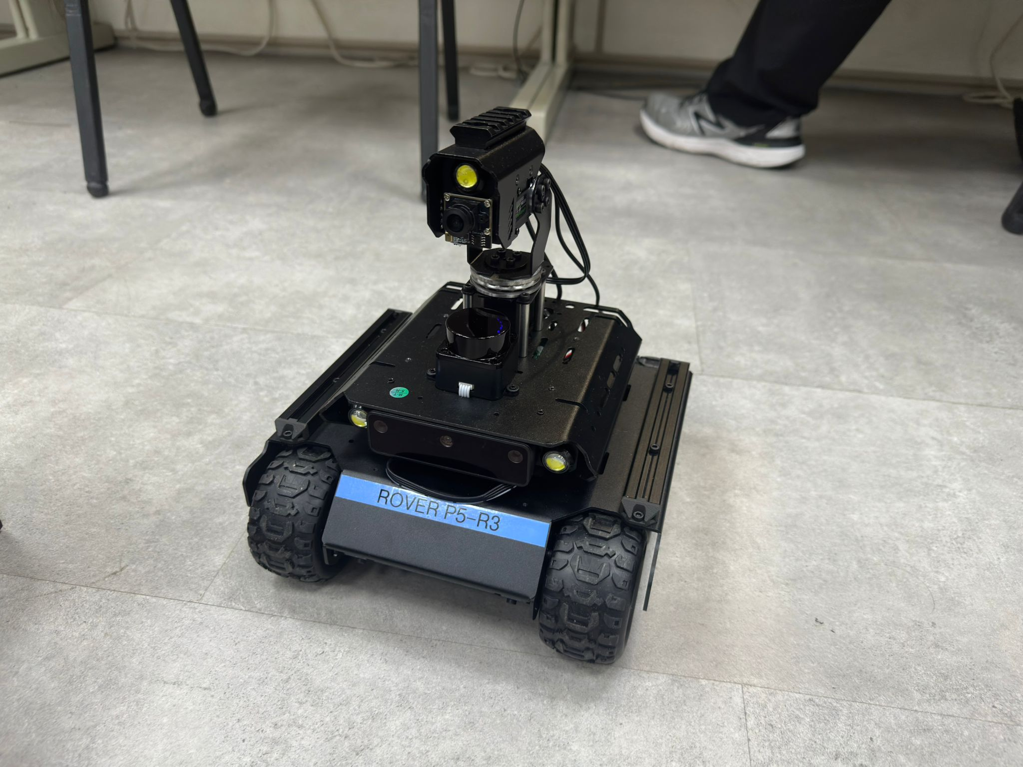 A mobile, autonomous robot prototype, designated ROVER P5-R3, resting on a light-colored floor. The robot has a sturdy black chassis and is equipped with four large wheels. A dedicated camera or sensor unit is mounted vertically on a mount at the front of the chassis.