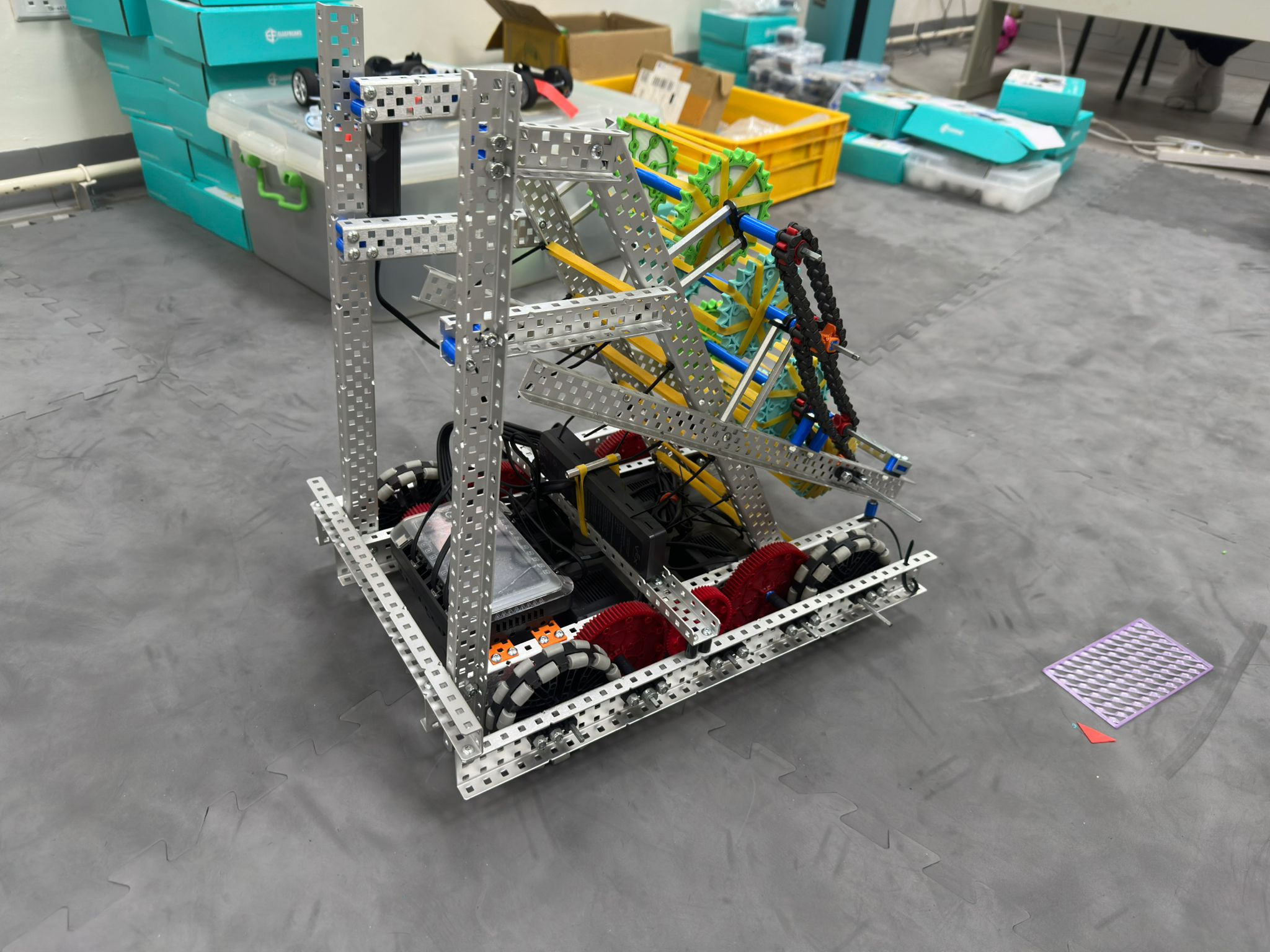 A large, complex, mechanical prototype structure built on the floor, assembled using numerous modular, silver metal rods and connectors. The robot appears to be highly articulated, featuring multiple vertical supports, linkages, and a base equipped with wheels, suggesting a sophisticated mechanical or lifting mechanism.